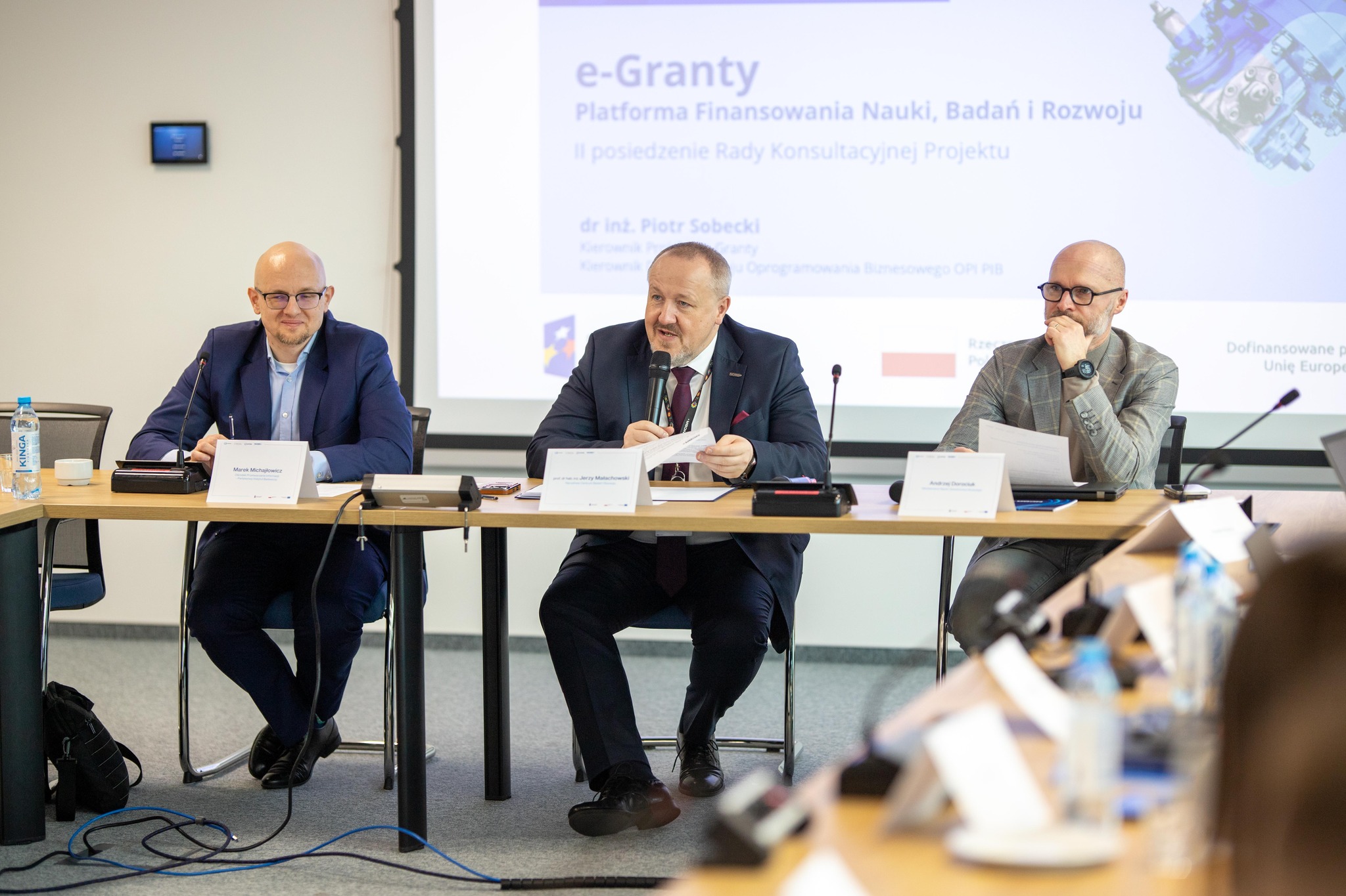 Trzech mężczyzn w strojach biznesowych siedzi za długim stołem podczas konferencji, prowadząc dyskusję. W tle widoczna prezentacja.